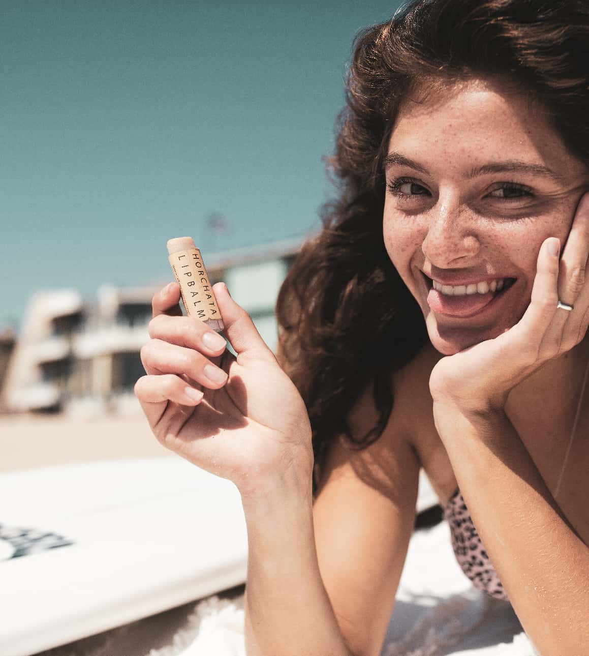 Woman holding SIN-MIN Horchata Lip Balm on the beach next to a surfboard
