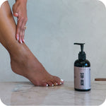 Person applying lotion to their leg with a bottle of 'SIN-MIN' lotion on a marble surface.