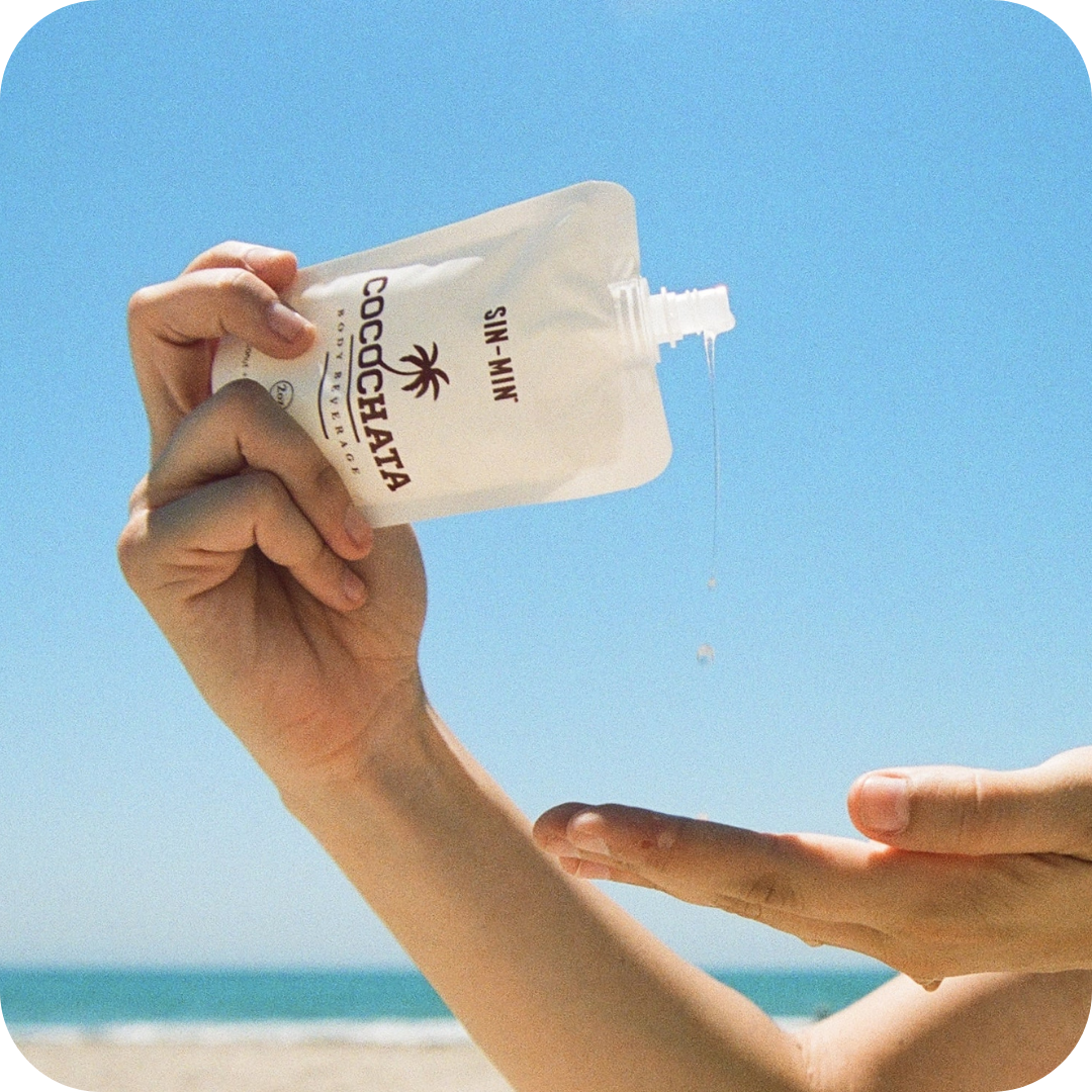 Hand applying Cocochata Body Beverage to another hand with a clear blue sky and beach in the background