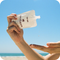 Hand applying Cocochata Body Beverage to another hand with a clear blue sky and beach in the background