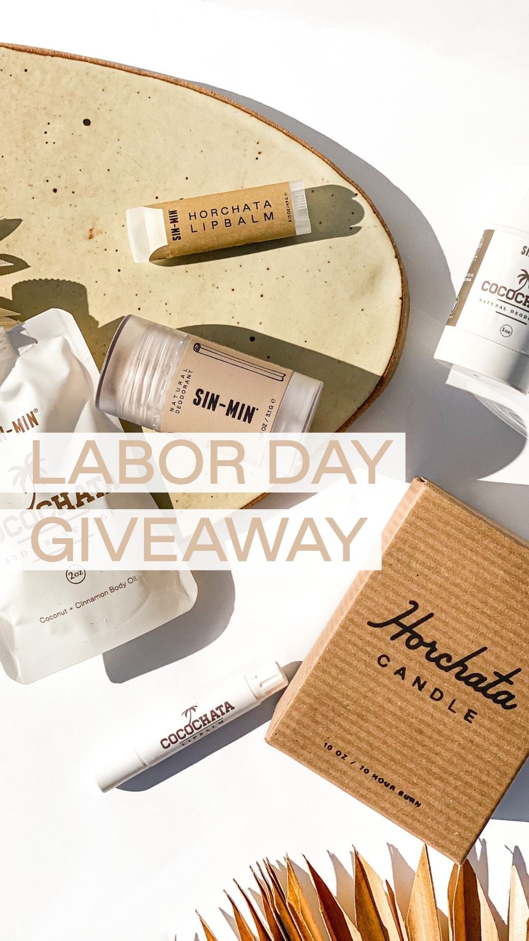 A Labor Day giveaway flat-lay featuring various Cosmatics products including a Horchata Lipbalm, Sin-Min body butter, and a Horchata Candle in its kraft paper box, arranged on a white surface with a beige ceramic plate and dried palm leaves.