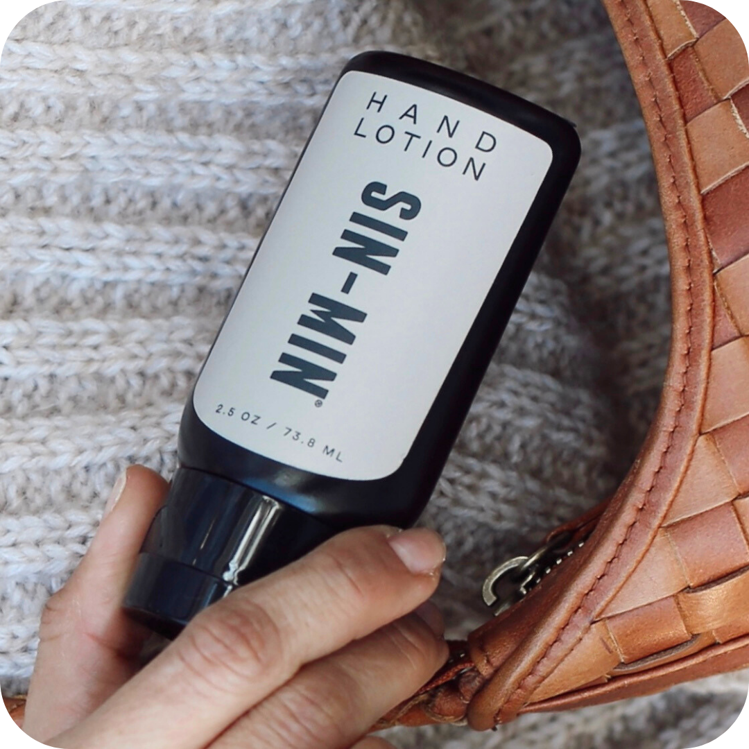 Hand holding a bottle of 'SIN-MIN' hand lotion next to a woven brown bag on a textured surface.