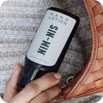 Hand holding a bottle of 'SIN-MIN' hand lotion next to a woven brown bag on a textured surface.
