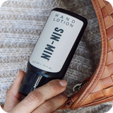 Hand holding a bottle of 'SIN-MIN' hand lotion next to a woven brown bag on a textured surface.