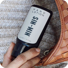 Hand holding a bottle of 'SIN-MIN' hand lotion next to a woven brown bag on a textured surface.