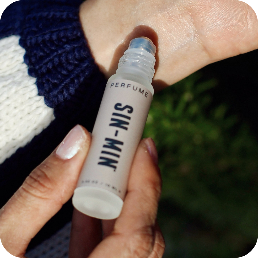 Hand holding a small perfume bottle labeled 'SIN-MIN' being applied to the inside of the other wrist.