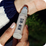 Hand holding a small perfume bottle labeled 'SIN-MIN' being applied to the inside of the other wrist.