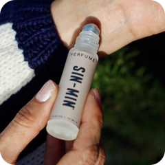 Hand holding a small perfume bottle labeled 'SIN-MIN' being applied to the inside of the other wrist.