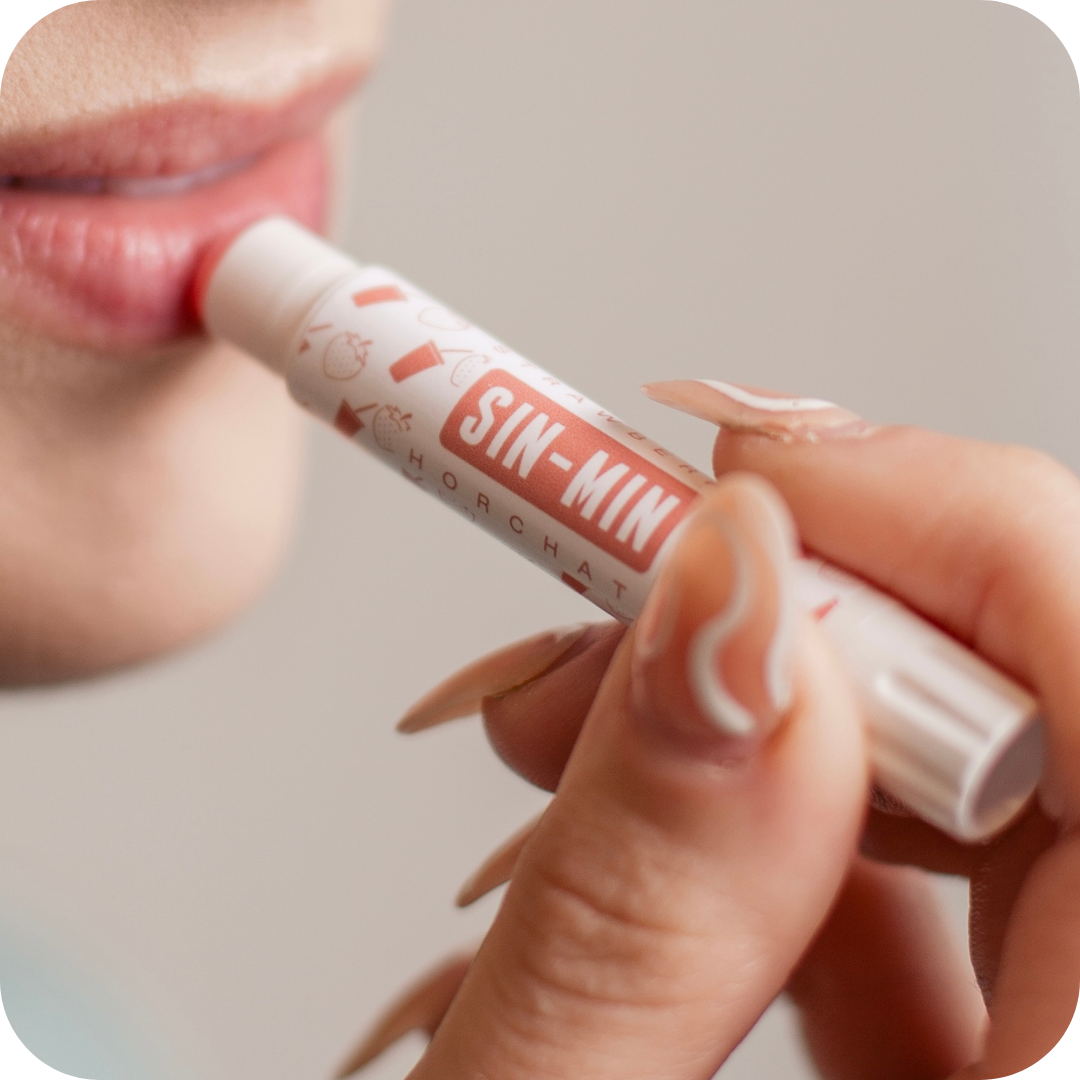 Person applying a lip balm labeled 'SIN-MIN' with a blurred background