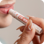 Person applying a lip balm labeled 'SIN-MIN' with a blurred background