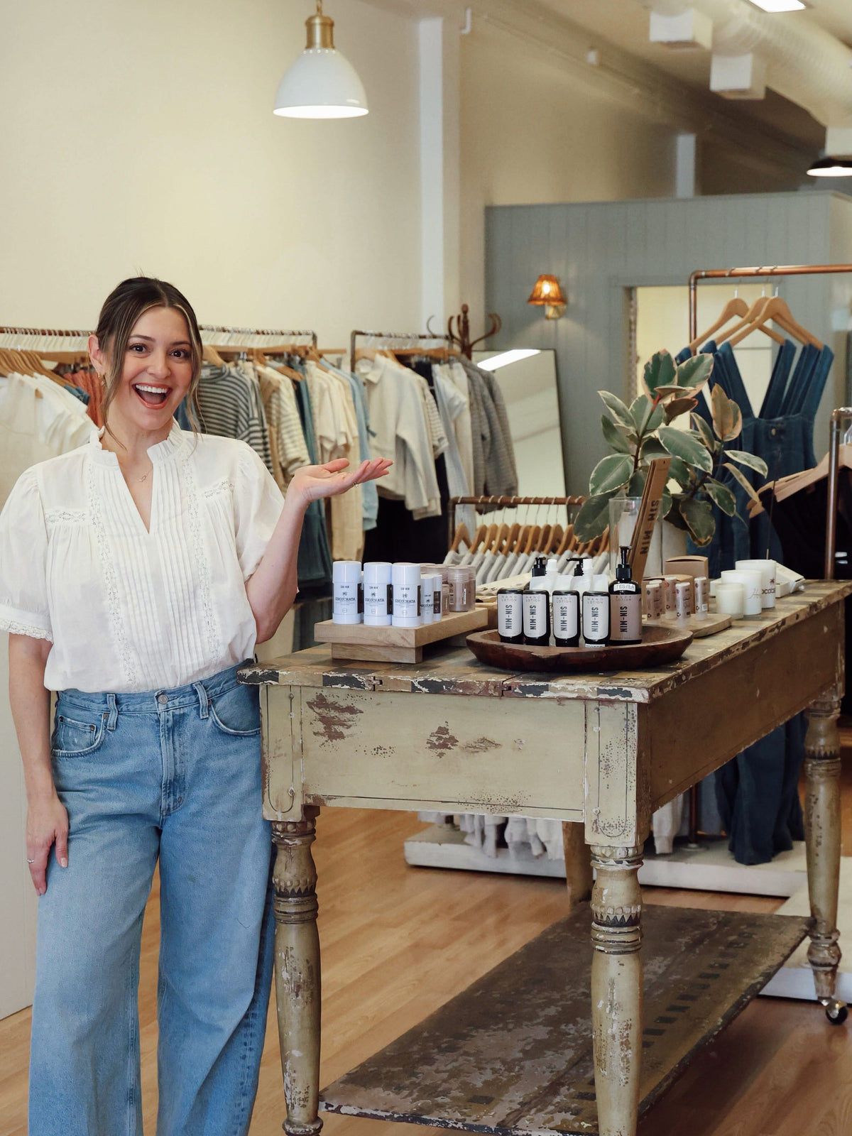 A woman smiling and gesturing towards a display table of skincare products inside a boutique. She is wearing a white pleated blouse and light blue jeans.